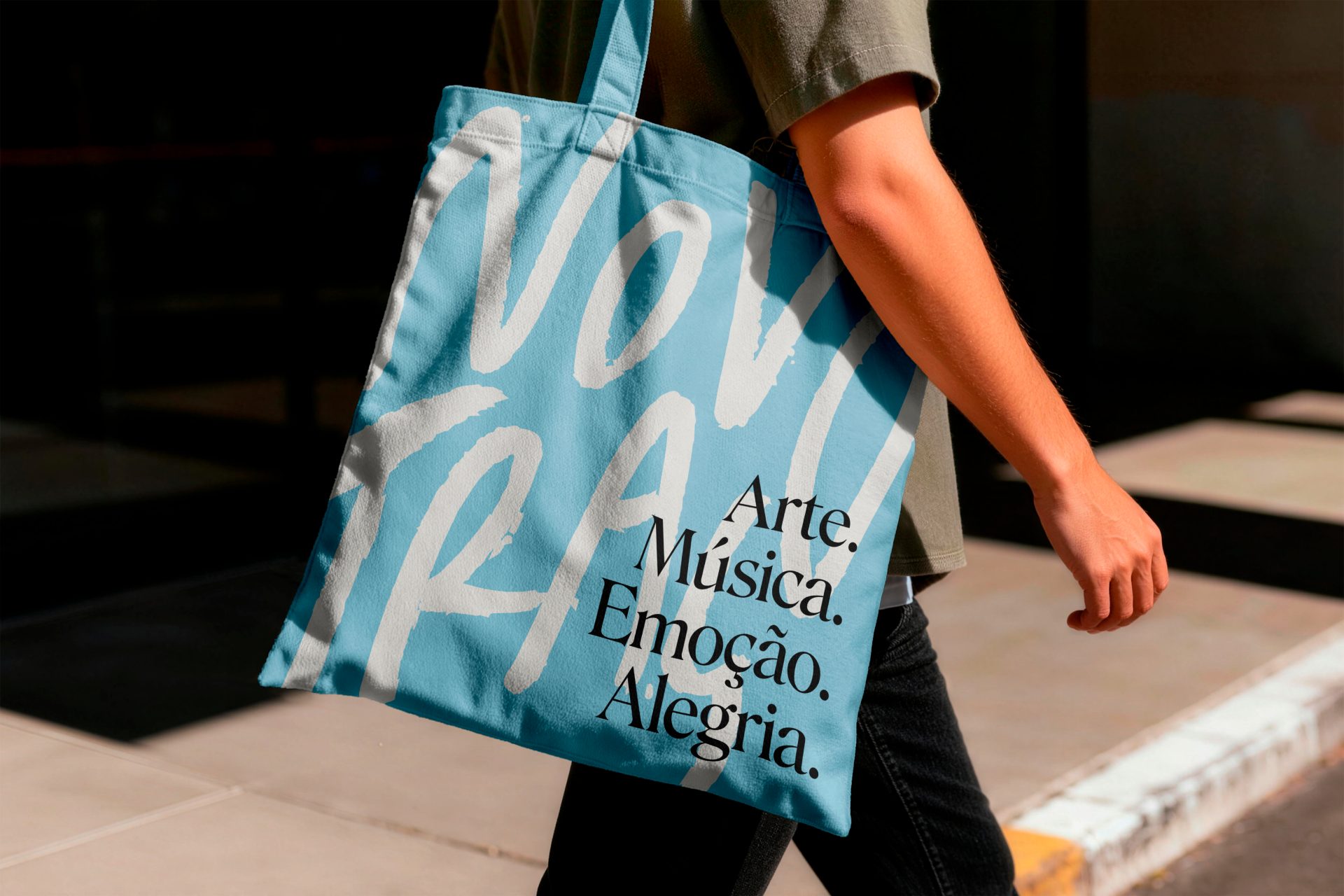 Pessoa caminhando com ecobag azul estampada com a assinatura manuscrita Novo Traço e as palavras Arte, Música, Emoção, Alegria