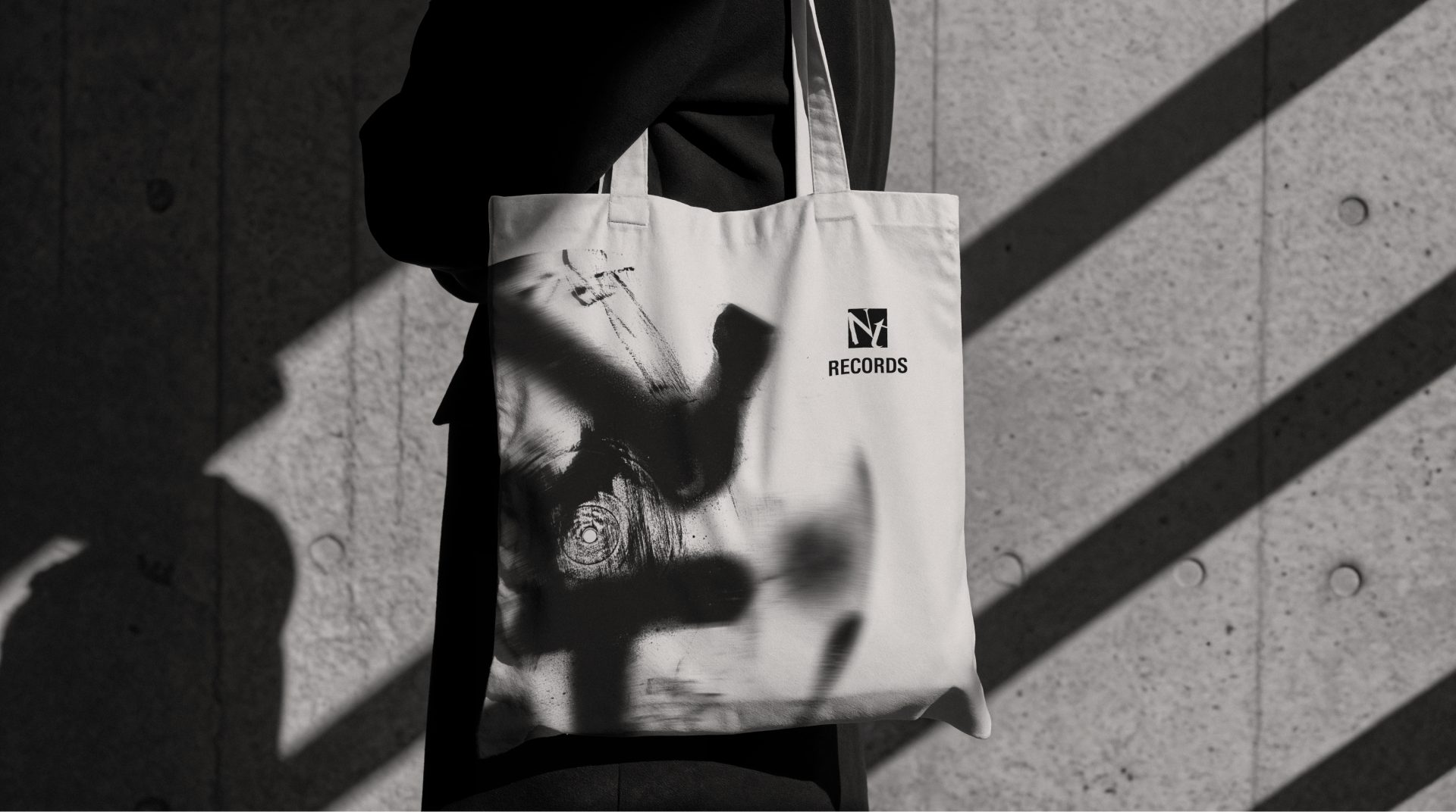 Ecobag creme com traços de spray em preto e o logo NT Records, carregada no ombro diante de uma parede de concreto com sombras diagonais