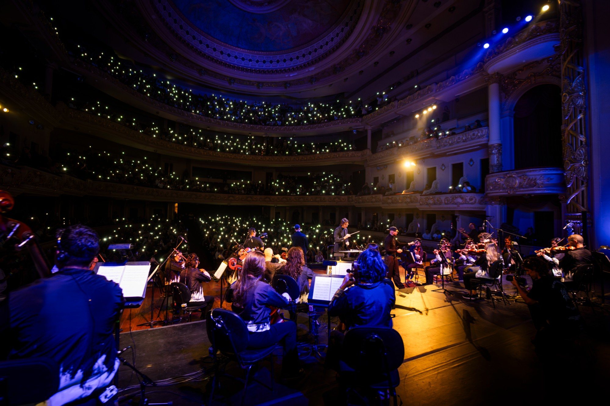 Vista do palco para a plateia: a orquestra tocando enquanto o público acende as lanternas dos celulares em todos os andares do teatro